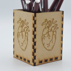 May include: A wooden pencil holder with a heart design. The holder is brown and has a black border. There are several pens in the holder.