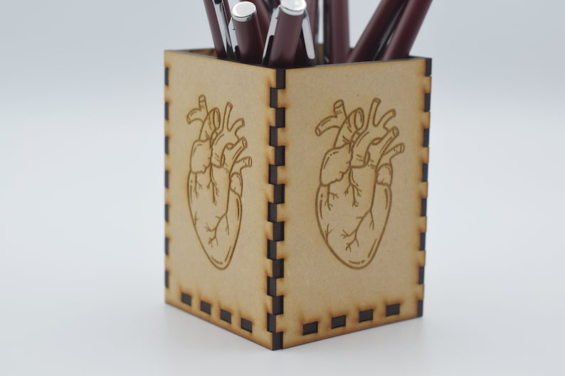 May include: A wooden pencil holder with a heart design. The holder is brown and has a black border. There are several pens in the holder.