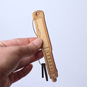 May include: A wooden ornament shaped like a syringe with a gold string attached to the top. The syringe has a brown wood grain finish and a black outline.