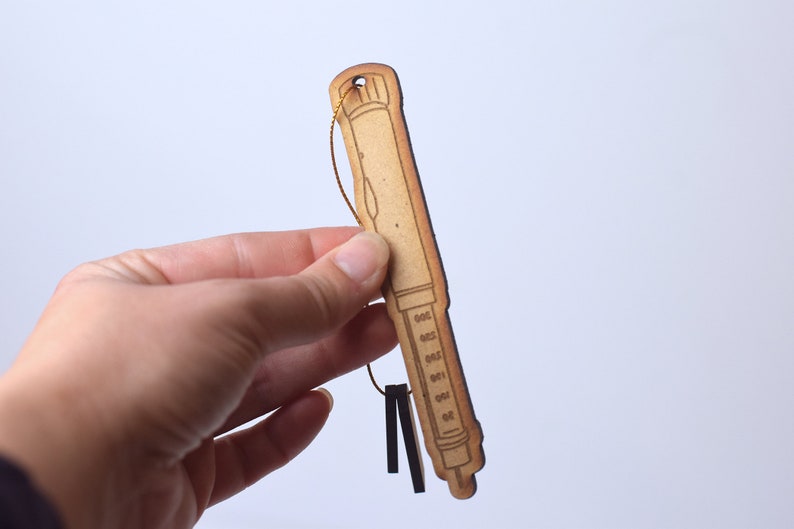 May include: A wooden ornament shaped like a syringe with a gold string attached to the top. The syringe has a brown wood grain finish and a black outline.
