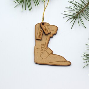 May include: Wooden Christmas ornament shaped like a ski boot with a brown finish. The ornament has two tags attached to it, one that says "YEAR" and the other that says "YOUR NAME".