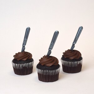May include: Three chocolate cupcakes with chocolate frosting and a silver plastic scalpel on top of each cupcake.