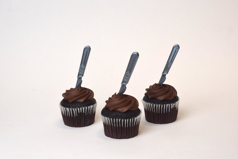 May include: Three chocolate cupcakes with chocolate frosting and a silver plastic scalpel on top of each cupcake.