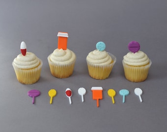 May include: Four cupcakes with vanilla frosting, each topped with a different-shaped, colorful pill or medicine bottle decoration. Below the cupcakes are several small, colorful, pill-shaped decorations.