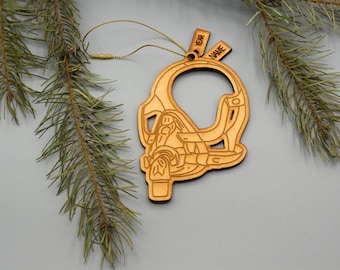 May include: A wooden Christmas ornament in the shape of a helmet, with the words "YEAR" and "NAME" on small tags. The ornament is light brown and hangs from a gold string, with pine branches in the background.