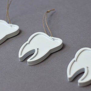 May include: Three white tooth-shaped ornaments with brown string hangers. The ornaments are arranged in a row on a gray surface.