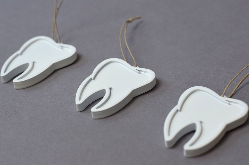 May include: Three white tooth-shaped ornaments with brown string hangers. The ornaments are arranged in a row on a gray surface.
