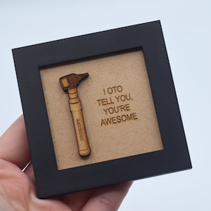 Op de afbeelding: Een klein, ingelijst houten bord met een bruine achtergrond. Het bord heeft een houten otoscoop en de tekst "I oto tell you, you're awesome".