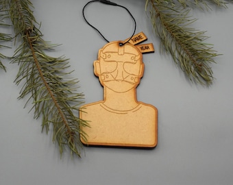 May include: A wooden ornament in the shape of a person wearing a mask, with the words "NAME" and "YEAR" on small tags. The ornament is light brown with a black string for hanging. Green pine branches are in the background.