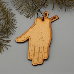 May include: A wooden hand-shaped ornament with a black outline and a string for hanging. Two small wooden tags are attached, one labeled "YEAR" and the other "NAME". The ornament is set against a grey background with pine branches.