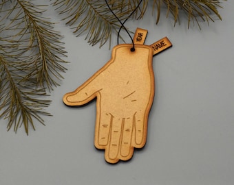 May include: A wooden hand-shaped ornament with a black outline and a string for hanging. Two small wooden tags are attached, one labeled "YEAR" and the other "NAME". The ornament is set against a gray background with pine branches.