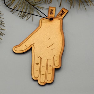May include: A wooden hand-shaped ornament with a black outline and a string for hanging. The ornament has three small rectangular tags that read "YEAR" and "NAME". The ornament is a light brown color.