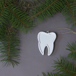 May include: White tooth-shaped Christmas ornament with a string loop for hanging. The ornament is surrounded by green pine branches on a gray background.