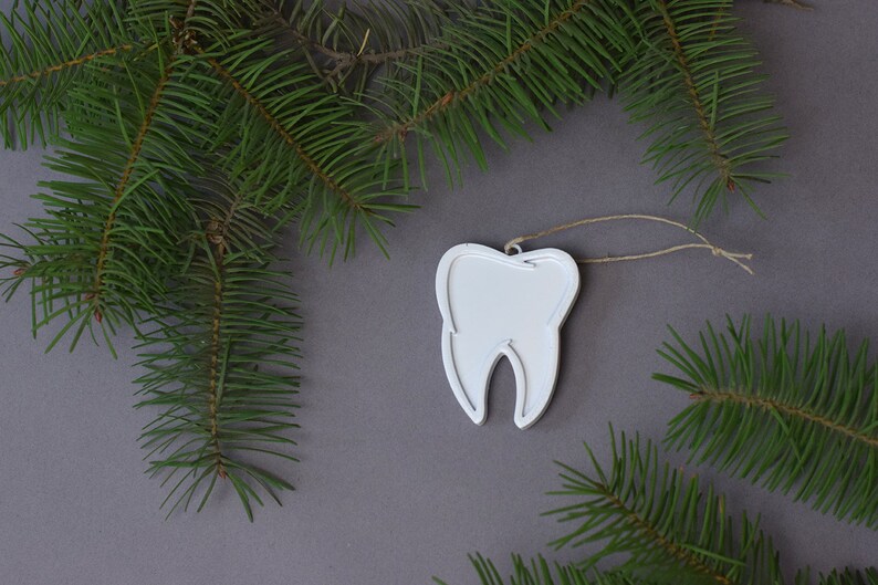 May include: White tooth-shaped Christmas ornament with a string loop for hanging. The ornament is surrounded by green pine branches on a gray background.