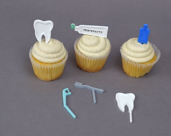 May include: Three cupcakes decorated with dental hygiene themed toppers. The cupcakes are topped with white frosting and sprinkles. The toppers include a tooth, a tube of toothpaste that says "Toothpaste", and a bottle of mouthwash. There are also two additional toppers: a toothbrush and a dental floss pick.