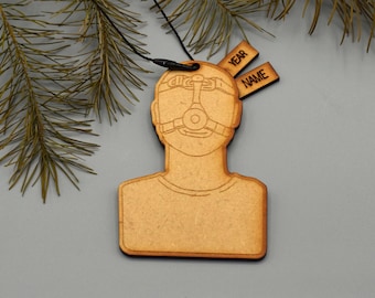May include: Wooden ornament in the shape of a person wearing a mask. The ornament has two small tags that read "YEAR" and "NAME". The ornament is hanging from a black string. The background includes pine needles.