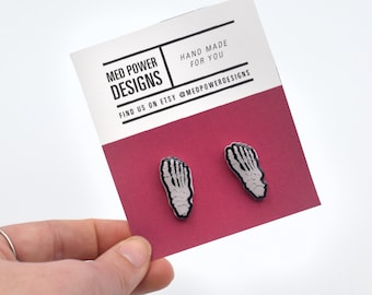 May include: Two white and black skeleton foot enamel pins on a red card with the text "MED POWER DESIGNS" and "HAND MADE FOR YOU" and "FIND US ON ETSY @MEDPOWERDESIGNS".