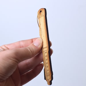 May include: A wooden ornament shaped like a syringe with a gold string attached to the top. The ornament is brown and has a black outline.