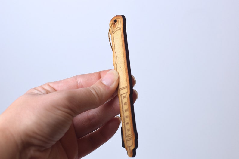 May include: A wooden ornament shaped like a syringe with a gold string attached to the top. The ornament is brown and has a black outline.