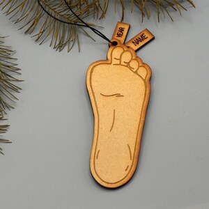 May include: Wooden foot-shaped ornament with the words "YEAR" and "NAME" on small tags. The ornament is light brown with a detailed outline of a foot. A black string is attached for hanging. The background is a light gray.