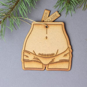 Puede incluir: Un adorno de madera con forma de vientre de embarazada con una línea cosida a través del abdomen. El adorno está decorado con un diseño simple de ropa interior. El adorno tiene una cuerda atada a la parte superior y dos etiquetas que dicen "YEAR" y "CLAIRE".
