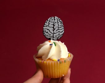 May include: A white cupcake with white icing and colourful sprinkles topped with a grey brain-shaped cake topper. The cupcake is held in a hand against a red background.