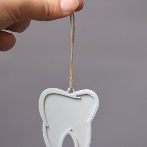 May include: A white tooth-shaped ornament with a string loop for hanging. The ornament is made of a smooth, white material and is shaped like a tooth.