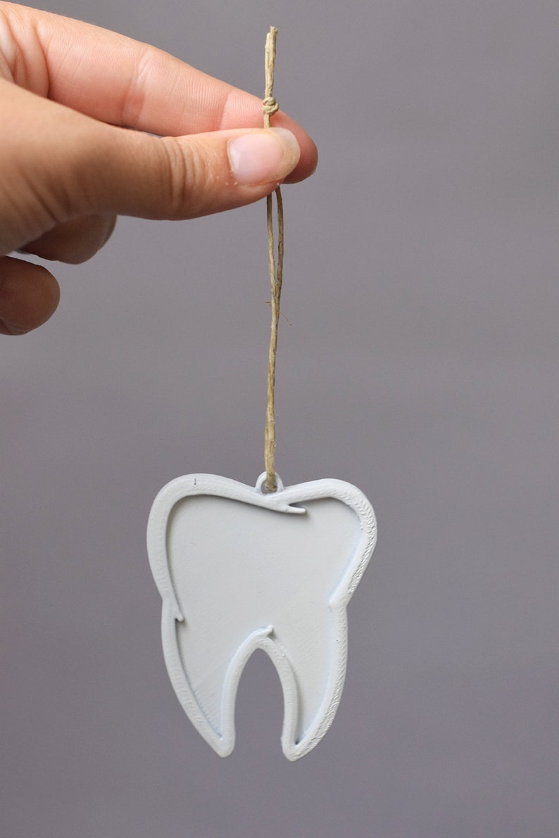 May include: A white tooth-shaped ornament with a string loop for hanging. The ornament is made of a smooth, white material and is shaped like a tooth.