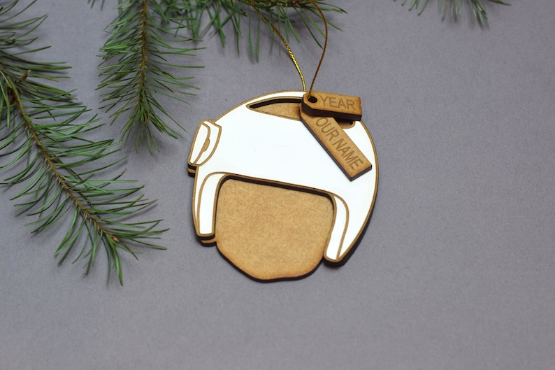 Pu&ograve; includere: Un ornamento natalizio in legno bianco a forma di casco con un'etichetta in legno marrone che dice "YEAR OUR NAME".