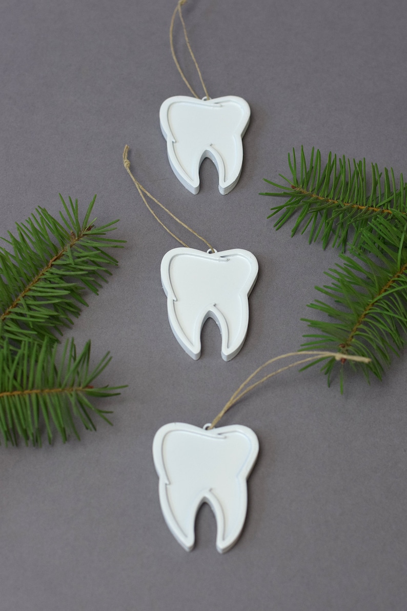 May include: Three white tooth-shaped ornaments with twine hangers. The ornaments are on a gray background with green pine branches.
