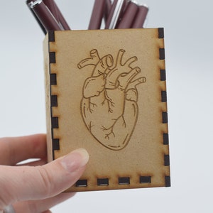 May include: A wooden pencil holder with a laser-engraved anatomical heart design. The holder is brown with black trim.
