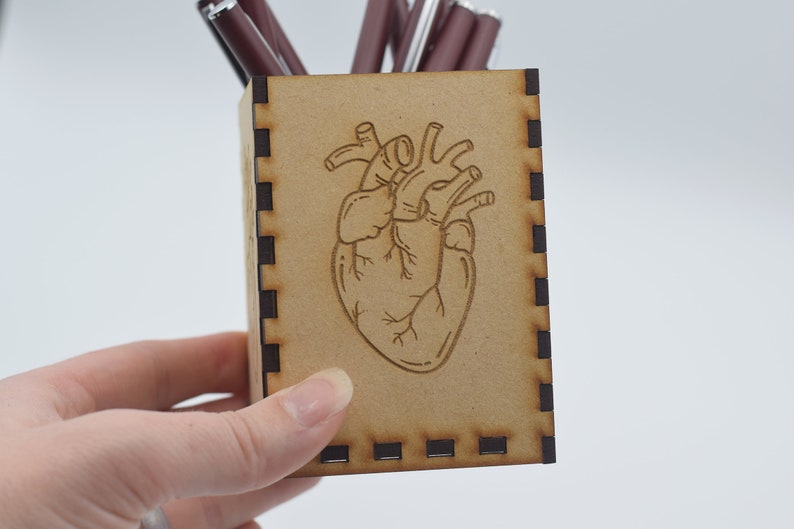 May include: A wooden pencil holder with a laser-engraved anatomical heart design. The holder is brown with black trim.