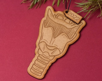 May include: A wooden Christmas ornament shaped like a Predator head with a 2022 tag. The ornament is brown and has a detailed design.