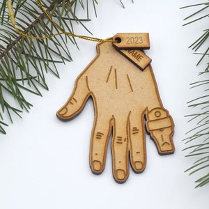May include: Wooden Christmas ornament shaped like a hand with a ring on the finger. The ornament has two tags attached to it, one with the year "2023" and the other with the word "NAME".