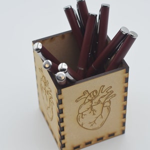 May include: A wooden pen holder with a heart design. The holder is filled with burgundy pens.