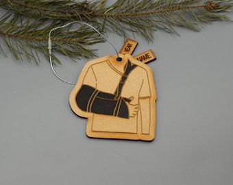 May include: Wooden Christmas ornament shaped like a person wearing an arm brace. The ornament is light brown with a black arm brace. The ornament has two small tags that say "YEAR" and "NAME". A silver string is attached for hanging.