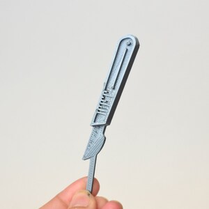 May include: A silver-colored scalpel with a handle and a blade. The scalpel is held in a hand.