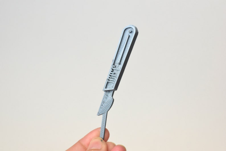 May include: A silver-colored scalpel with a handle and a blade. The scalpel is held in a hand.