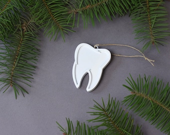 May include: White tooth-shaped Christmas ornament with a twine string hanging from a gray background with green pine branches.