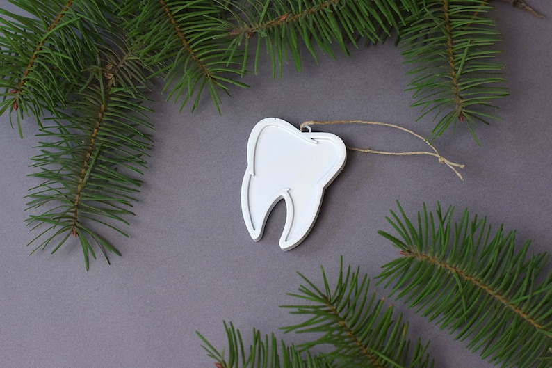 May include: White tooth-shaped Christmas ornament with a twine string hanging from a gray background with green pine branches.