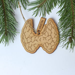 May include: A wooden thyroid ornament with the name "Kristin" and the word "Thyroid" engraved on a small tag. The ornament is light brown and hangs from a string, with a detailed anatomical design. Green pine branches are in the background.