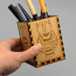 May include: A wooden pen holder with a laser-etched anatomical design. The holder is square-shaped and contains several pens and markers. The design appears to be a stylized representation of a thyroid gland. The holder is held by a person.