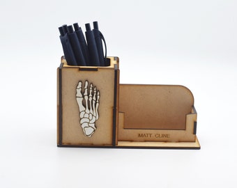 May include: Wooden desk organizer with a skeleton foot design and a separate compartment for business cards. The compartment has the text "MATT. CLINE" engraved on it.