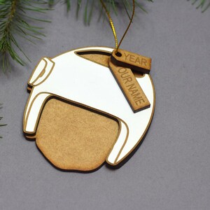 Pu&ograve; includere: Un ornamento natalizio in legno a forma di casco con un design dipinto di bianco. L'ornamento ha un'etichetta attaccata con il testo "YEAR" e "OUR NAME".