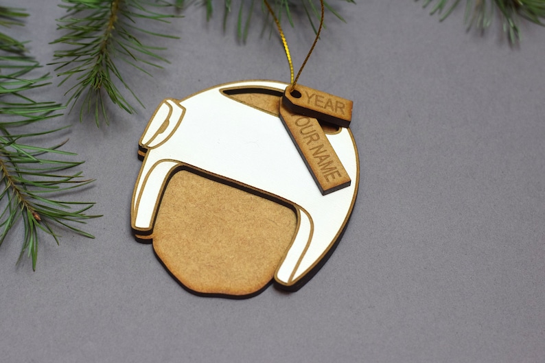 Pu&ograve; includere: Un ornamento natalizio in legno a forma di casco con un design dipinto di bianco. L'ornamento ha un'etichetta attaccata con il testo "YEAR" e "OUR NAME".
