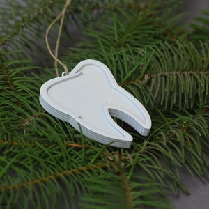 May include: White tooth-shaped Christmas ornament hanging from a pine branch. The ornament is made of layered wood and has a rustic, handmade look.
