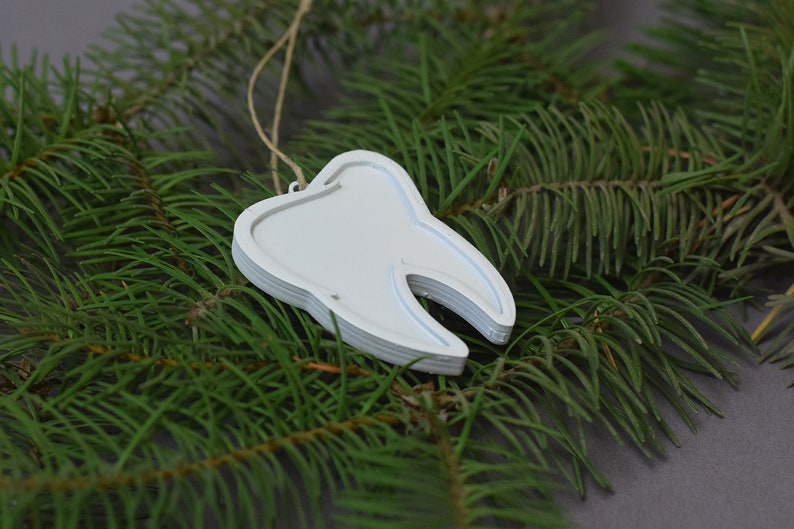 May include: White tooth-shaped Christmas ornament hanging from a pine branch. The ornament is made of layered wood and has a rustic, handmade look.