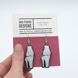 May include: A pair of silver dangle earrings featuring a white anatomical knee bone design. The earrings are displayed on a card with the text "Med Power Designs" and "Hand Made For You".