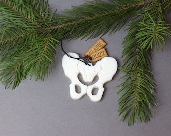 May include: White ceramic ornament shaped like a human pelvis with a black string for hanging. Two brown wooden tags are attached to the ornament, one says "Claire" and the other says "2022".