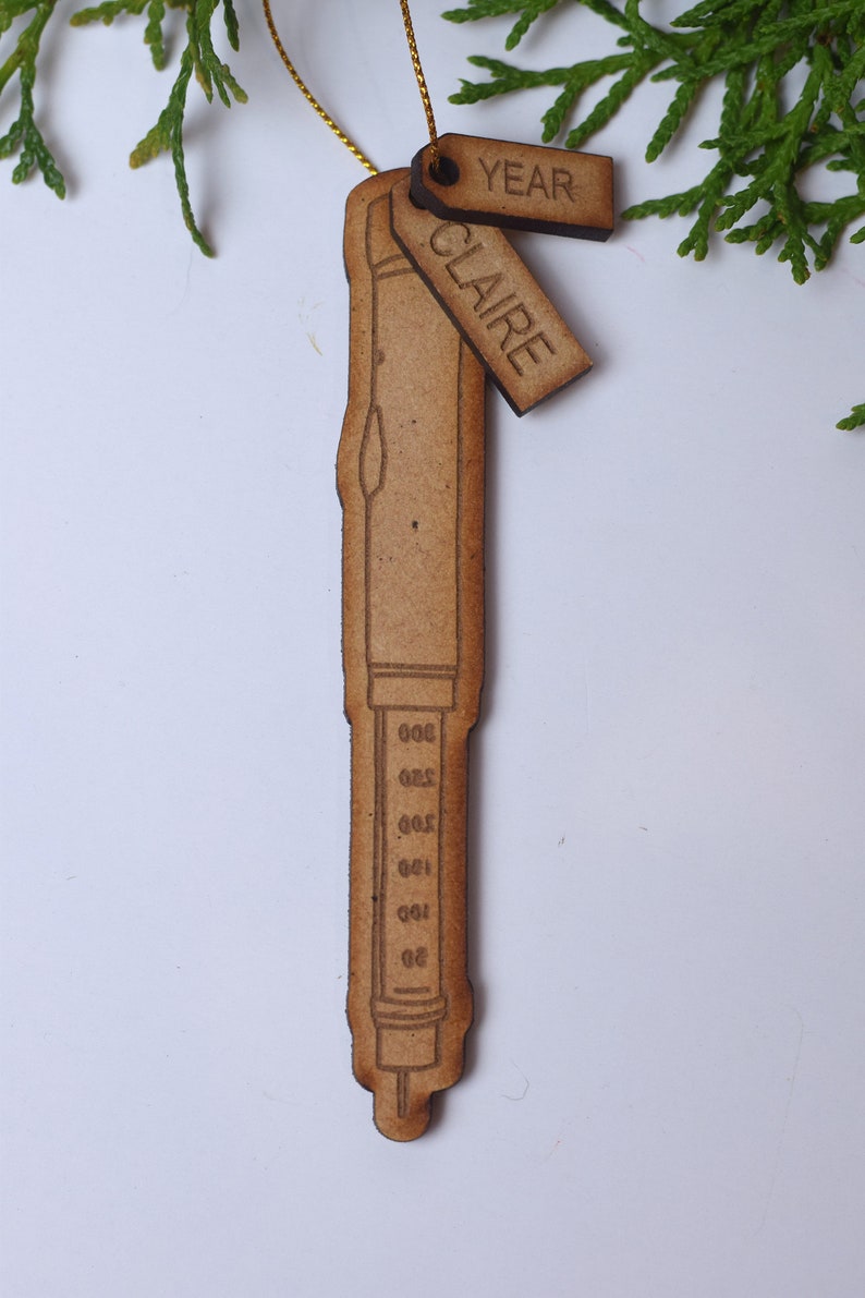 May include: A wooden ornament shaped like a syringe with a scale on the side. The ornament has two tags attached to it, one that says "YEAR" and another that says "CLAIRE".